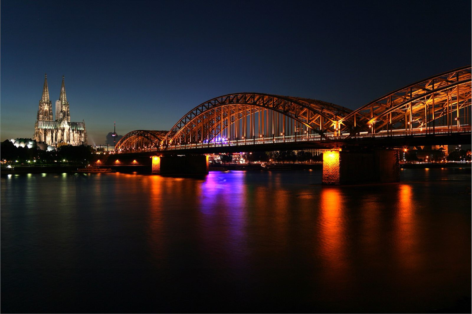 The Hohenzollern Bridge and Cologne Cathedral lit at night, reflected in the Rhine.