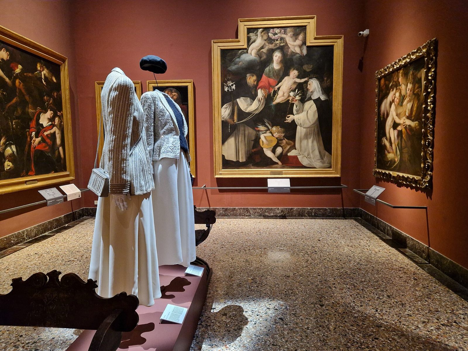 A red-walled room of the Pinacoteca di Brera, with altarpieces and two gowns on display.