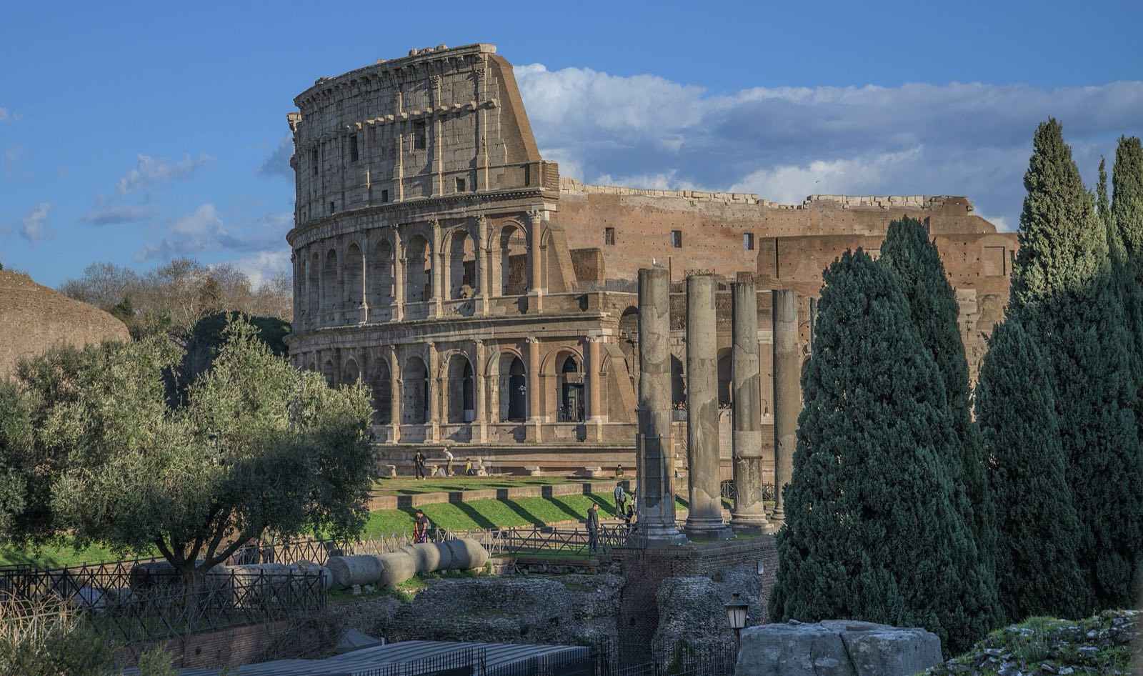 The Colosseum seen from the Roman Forum, behind ancient columns and cypress trees.